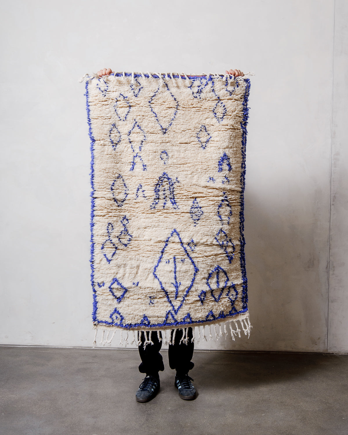 Berberlin Azilal rug in cream wool with blue cotton design being held up in front of a white wall.